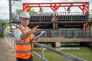 an engineer in safety vest inspects water gate structure, recording data on a sunny day