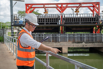 an engineer in safety vest inspects water gate structure, recording data on a sunny day