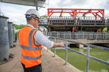 an engineer in safety vest inspects water gate structure, recording data on a sunny day