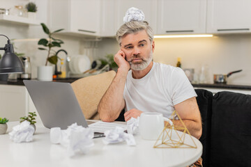 Middle-aged man at home writes ideas for project at table with laptop and crumpled paper feels stuck and tired. Mature guy battles creative block shows frustration doubt seeks focus to continue work.