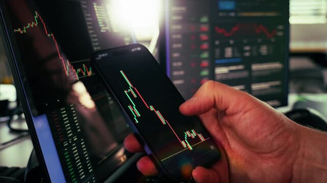 Cryptocurrency market volatility mobile analysis trading screens. Close-up of a hand holding smartphone showing candlestick chart with red and green indicators, in front of trading monitors with finan