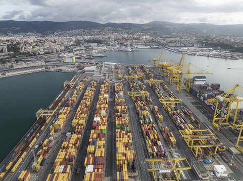 Trieste, Italy - 10 October 2025: Aerial view of a bustling port where rows of colorful shipping containers meet the Adriatic Sea, framed by the city's skyline and distant mountains.
