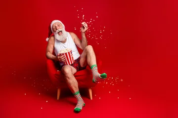 Fotobehang Beren Santa enjoys popcorn on Christmas Eve relaxing in a red chair wearing hat striped socks and smiling  © deagreez