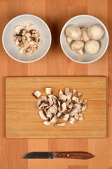 An overhead shot of fresh whole and chopped white button mushrooms in white bowls and on a wooden cutting board with a knife