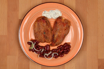 An overhead shot of a plate with breaded roasted stuffed red peppers served with a dollop of tartar sauce and a side of bean salad on a wooden background