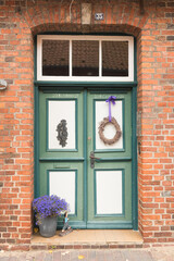 Vintage green wooden door with white panels set in a rustic brick wall. A wreath with a purple ribbon decorates the entrance, and a pot of violet flowers adds charm and warmth to the cozy doorway