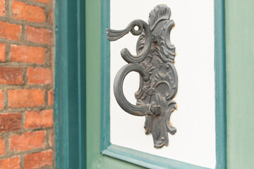 A detailed close-up of an ornate, antique black metal door pull featuring a ring and scrollwork, mounted on a white door panel framed by teal trim. A red brick wall is visible on the left