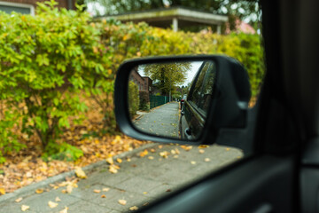 Close-up view of a car side mirror reflecting a quiet residential street with green bushes and fallen autumn leaves. The image conveys a sense of travel, observation, and everyday road