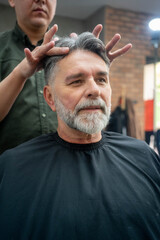 Fototapeta premium Barber’s hands styling mature man’s hair as client admires haircut at salon