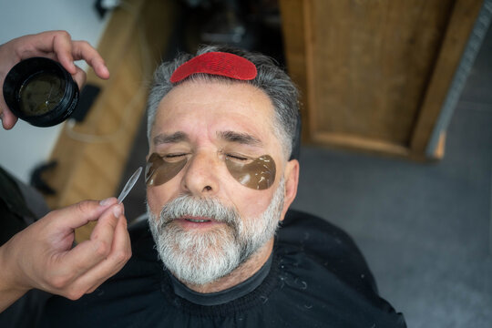 Mature man receiving under-eye gel patches as part of luxurious barber facial grooming treatment