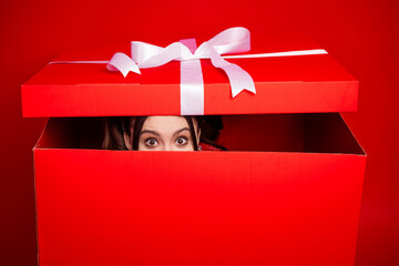 A surprised woman peeks from a large red gift box with a white ribbon in a festive christmas scene perfect for holiday shopping and gift giving