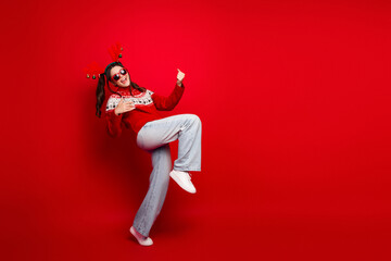 Young woman in red Christmas sweater with reindeer antlers poses playfully against a bright red...