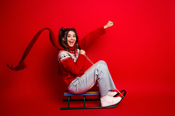 Joyful young woman on a sled in a red sweater poses playfully against a bright red background for a...