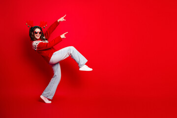 Joyful woman in red sweater dancing against red background with christmas antlers and sunglasses in...
