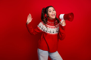 Cheerful young woman in red sweater with megaphone posing against bold red background for festive...