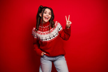 Happy young woman in red Christmas sweater smiles and shows peace sign against red background for...