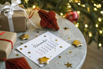 December calendar surrounded by Christmas gifts and red Christmas tree decorations on marble table near Christmas tree