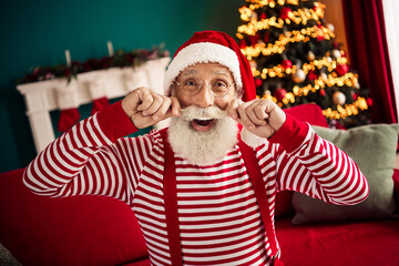 Fototapeta premium Joyful elderly man dressed as Santa Claus posing playfully by a Christmas tree in a cozy vibrant room