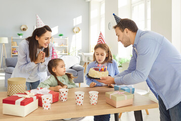 Joyful family celebrating child birthday at home, son making wish and blowing candles. Happy parents and kids wearing party hats enjoying festive atmosphere with gifts and cake. Warm family moment.