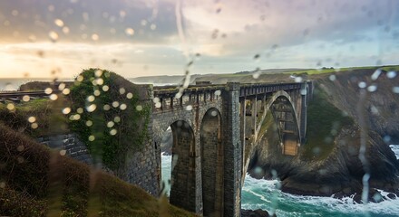 Rain-Streaked Window View of Historic Stone Viaduct Spanning Rugged Coastal Cliffs and Crashing Waves at Sunset.