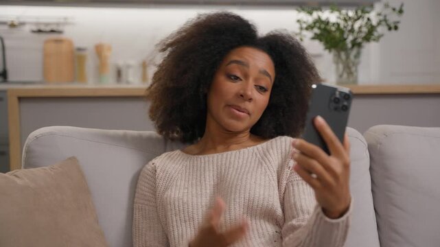 Positive African American woman talking mobile phone video call virtual chat influencer vlogger talk smartphone online conference in living room at home girl female greeting hello remote communication