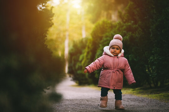 A beautiful little girl in pink warm clothes is walking in a park at sunset.
