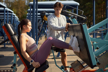 Caucasian young adult woman exercising on leg press machine outdoors while Caucasian young adult man supervising workout, both focused on fitness training in open air gym setting
