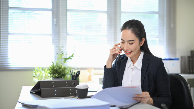Fototapeta Businesswoman talking on the phone while reviewing documents at her desk, showing client consultation
