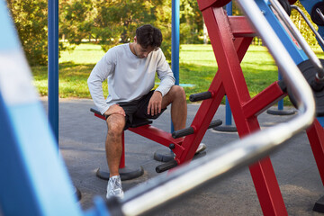Young adult Caucasian man sitting on outdoor gym bench resting after workout, looking down with hand on thigh, exercise equipment surrounding, park setting visible in background © DragonImages
