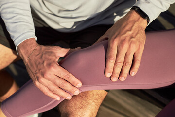 Caucasian young adult man massaging knee of Caucasian young adult woman outdoors, hands gently pressing on leg, both wearing athletic clothing, close up on interaction
