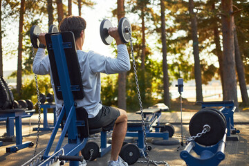 Caucasian young adult man performing shoulder press exercise on outdoor gym equipment, lifting dumbbells with chains attached, surrounded by weightlifting machines in forest setting