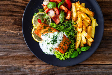 Fried salmon steak with tzatziki sauce, homemade potato fries and tomato salad on wooden table. Top view