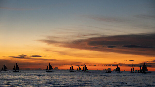 Multiple sailboats silhouetted against a vivid orange and blue sunset sky over a calm tropical sea