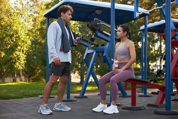 Caucasian young adult man standing and holding smartphone while talking to Caucasian young adult woman sitting on outdoor gym bench, both wearing sportswear, fitness equipment visible