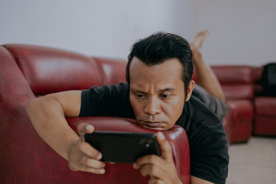 Young asian man using smartphone while lying relax on the couch enjoying video content in living room at home.