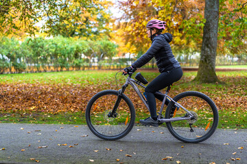Naklejka premium Middle aged beautiful woman riding bicycle in city park in autumn. Side view 