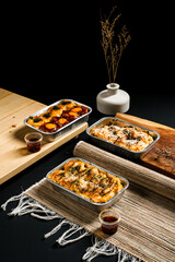 Flavor variants of baked dimsum served in aluminum trays — cheese, spicy red sauce, and sesame topping. Asian street food concept with minimal dark background.