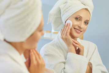 Blonde woman using cotton pad to apply toner and cleanse skin, practice skincare facial beauty routine at home. Young lady in bathrobe and towel on hair smiling at reflection in mirror, cleansing face