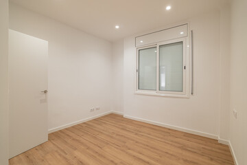 Empty room with white walls and wooden floor. A window allows natural light to enter. The door is closed, creating a minimalist and spacious atmosphere.