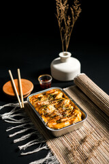 Side view of baked cheese dimsum in aluminum tray with creamy topping and herbs. Modern Asian comfort food photography on minimal dark background.