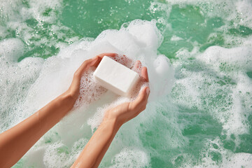 Hands hold a bar of white soap over foamy water in the bath