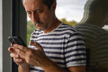 Mature man in striped sailor shirt standing on balcony and texting on smartphone, reflected in window glass, symbolizing modern communication and solitude.