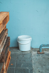 White plastic bucket on concrete floor beside stacked wooden pallets and blue wall. Minimal industrial scene with white bucket, blue wall, and weathered metal pipe detail.