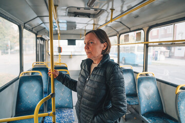 Casual mature woman standing inside old worn public bus, holding grip bar near exit door, empty interior of Eastern European city transport.