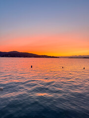 A serene sunset over a calm lake. The sky displays vibrant hues of orange, pink, and purple. Silhouettes of distant hills are visible on the horizon.