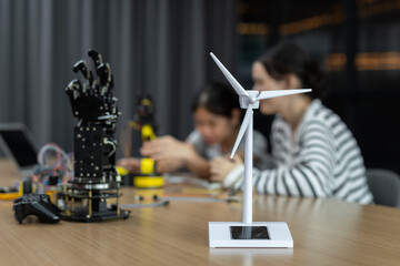 Close-up of wind turbine model with blurred girls working on robotics in background, representing renewable energy, STEM education and future technology innovation.