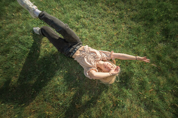 Young woman with blonde hair lies on green grass, wearing casual clothing, enjoying a sunny day...