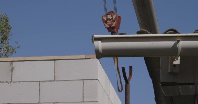 Industrial crane hook carefully lowering a heavy wooden rafter for roof construction as a skilled builder guides it into place on top of a concrete block wall against a clear blue sky.