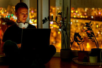 A young male developer is programming a code script late at night with beautiful city lights at night.