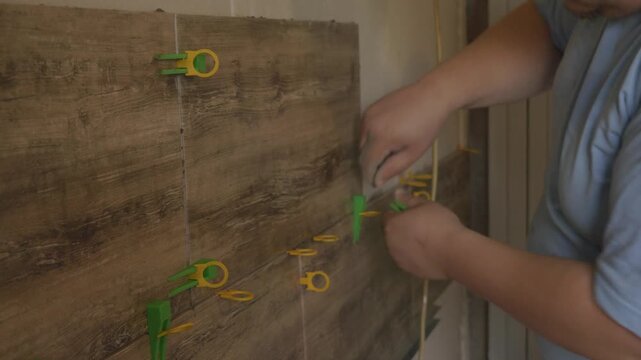 A professional tiler carefully lays large-format ceramic tiles on a wall. The tiler uses a leveling system with plastic clips and wedges to precisely install the tiles during a kitchen renovation.